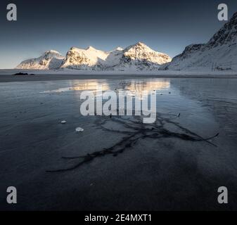 Coucher de soleil d'hiver sur la plage de Skagsanden, avec des textures intéressantes dans la glace en miroir reflétant les montagnes lumineuses en arrière-plan. Banque D'Images