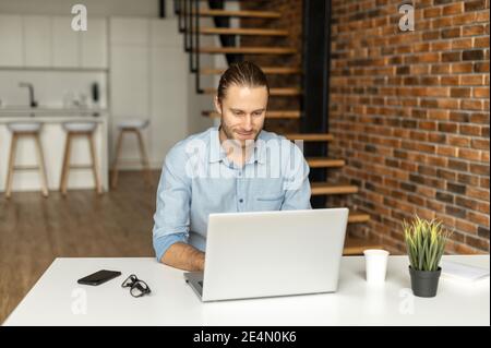 Un travail de vêtements hipster souriants à domicile. Un beau jeune entrepreneur dans le port chic décontracté utilisant un ordinateur portable pour la communication en ligne avec les clients et Banque D'Images