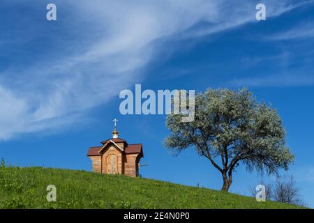 Petite ancienne chapelle en bois sur le sommet d'une colline herbeuse vert d'été, saule solitaire et ciel bleu avec nuage. Banque D'Images