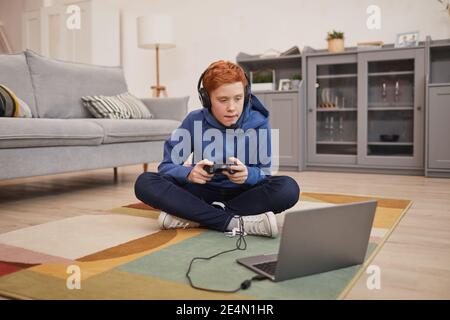 Portrait complet d'un adolescent aux cheveux rouges jouant à des jeux vidéo tout en étant assis sur le sol et tenant une manette de jeu, espace de copie Banque D'Images