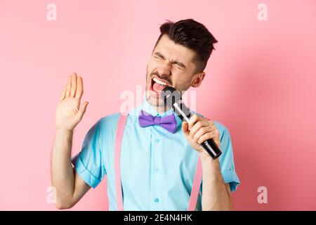 Concept de fête et d'événements festifs. Un gars drôle interprète une chanson, chantant le karaoké dans le microphone avec le visage insouciant, debout dans le nœud papillon et les bretelles sur Banque D'Images