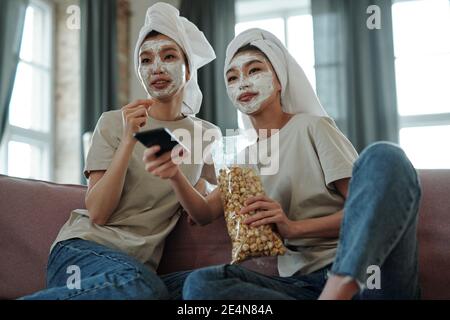 Jeunes femmes asiatiques heureuses avec des serviettes sur la tête et un masque en argile sur les visages assis devant le téléviseur, regarder le film et avoir du pop-corn Banque D'Images