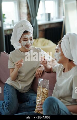 Jeune femme décontractée jumeaux avec des serviettes sur la tête et un masque en argile sur les visages assis sur le canapé à la maison, discutant de choses curieuses et ayant du pop-corn Banque D'Images