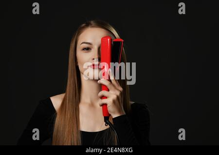 Jeune souriante belle femme avec de longs cheveux lisses soyeux dedans des vêtements noirs tenant le lisseur rouge à la main au-dessus d'un œil Banque D'Images