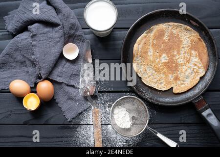 Crêpe chaude dans une casserole noire sur une table noire avec farine, lait et œufs. Banque D'Images