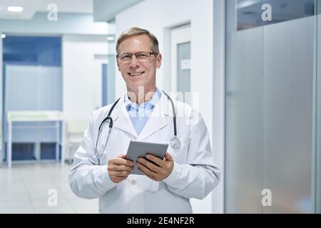 Un vieux médecin souriant d'âge moyen utilisant une tablette numérique à l'hôpital. Banque D'Images