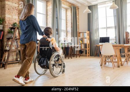 Vue arrière d'une jeune femme poussant un fauteuil roulant avec sa désactivation collègue ou ami tout en prenant soin d'elle et en aidant pour se déplacer dans la maison Banque D'Images