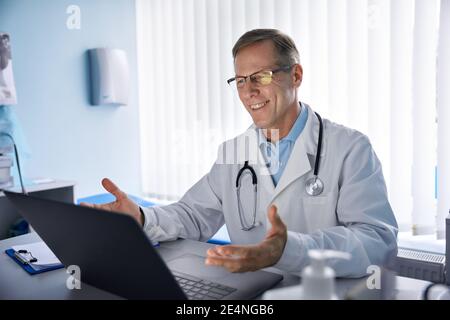 Bon vieux médecin de sexe masculin consultant le patient en ligne par appel vidéo sur ordinateur portable. Banque D'Images
