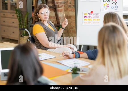 Groupe de jeunes femmes designers assis devant leur collègue en fauteuil roulant faisant la présentation de nouvelles idées sur tableau blanc Banque D'Images