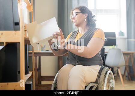 Jeune femme de bureau désactivée regardant à travers les papiers dans un seul de dossiers avec des documents sur l'étagère pendant la recherche de contrat exemple Banque D'Images