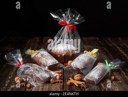 Le pain de Noël stollen, dans un emballage plastique et un ruban, est posé sur une planche et une table en bois, avec des noix et des épices. Banque D'Images