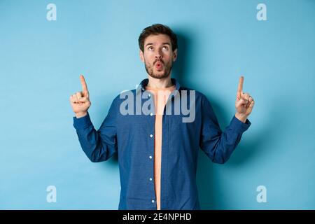 Image d'un jeune homme surpris qui dit wow, regardant et pointant les doigts vers le haut à l'offre promo, debout sur fond bleu Banque D'Images