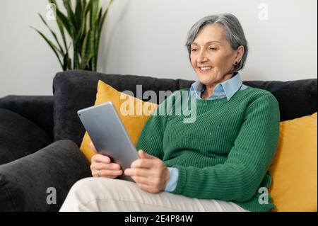 Une dame sénior dans un chandail vert, confortablement assise sur le canapé avec des oreillers jaunes dans le salon, regardant le spectacle de comédie sur le Banque D'Images