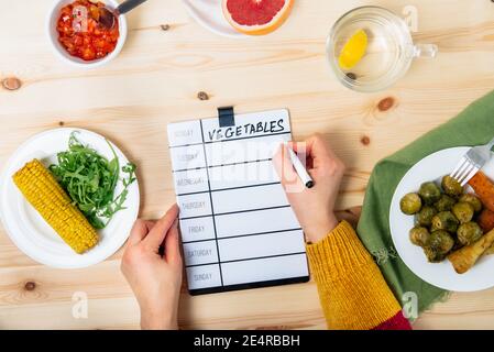 Planification du régime alimentaire et repas santé. Vue de dessus une femme écrivant à bord un menu de légumes hebdomadaire. Portion repas prête de légumes cuits sur la table en bois. Nouveau Banque D'Images