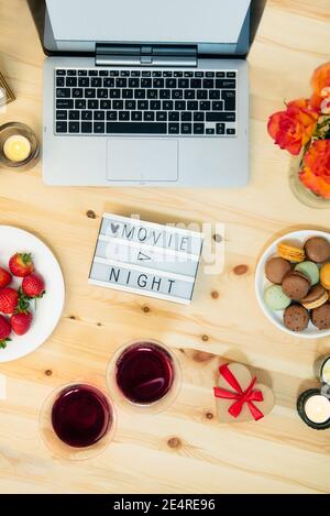 Vue de dessus romantique film Night concept. Message de nuit de cinéma sur un tableau lumineux, un ordinateur portable, des bougies, des fleurs, des macarons, des baies et deux verres avec vin pour Banque D'Images