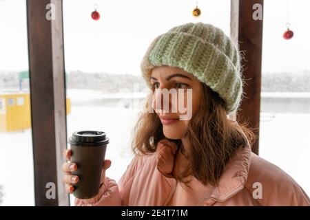 Ravie positive fille tenant une tasse de café en papier noir, souriant tendinement, debout seul dans le café, ayant le café dans sa main, étant de bonne humeur Banque D'Images