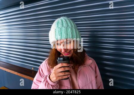 Ravie positive fille tenant une tasse de café en papier noir, souriant tendinement, debout seul dans le café, ayant le café dans sa main, étant de bonne humeur Banque D'Images