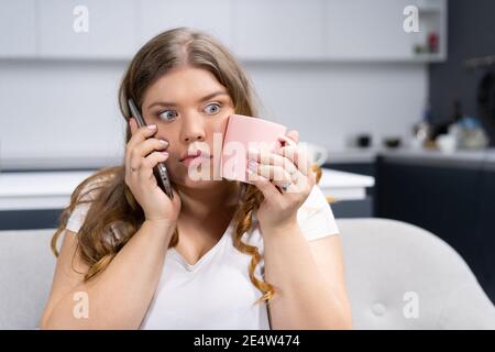 Choqué ou surpris look surpoids jeune femme parlant au téléphone avec un patron ou des amis tenant une tasse avec de l'eau froide touchant la poussin. Élégant Banque D'Images