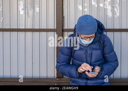 Un homme mature dans un masque médical s'assoit à un arrêt de bus à l'automne et attend un bus. Il recherche des informations sur l'emplacement des transports en commun. Banque D'Images