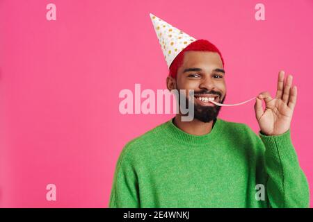 Joyeux afro-américain gars dans le cône de partie faire du plaisir avec chewing-gum isolé sur fond rose Banque D'Images