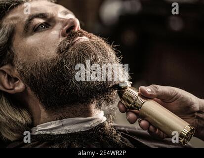 Homme barbu dans un salon de coiffure. Homme en visite chez le coiffeur dans le salon de coiffure. Le coiffeur fonctionne avec une tondeuse à barbe. Client Hipster qui obtient une coupe de cheveux. Mains d'un Banque D'Images