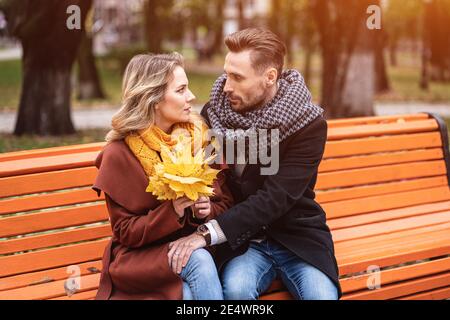 Couple amoureux heureux embrassant assis sur le banc romantique embrassé dans le parc portant des manteaux et des foulards collectant un bouquet de feuilles mortes. Histoire d'amour Banque D'Images