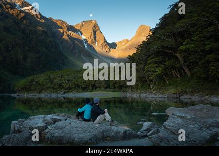 Un couple assis sur les rochers et appréciant la vue sur le lac Mackenzie à Routeburn Track dans South Island. Lever de lune et coucher de soleil en même temps. Banque D'Images