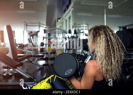 Femme aux cheveux longs et blonds bouclés tient une haltère lourde dans ses mains. Culturisme, entraîneur du club de fitness, salle de sport, vêtements de sport pour trai Banque D'Images