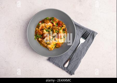 Poisson sicilien traditionnel avec tomates, olives, persil et poivron rouge dans une assiette profonde avec serviette et fourchette sur fond clair, gros plan Banque D'Images