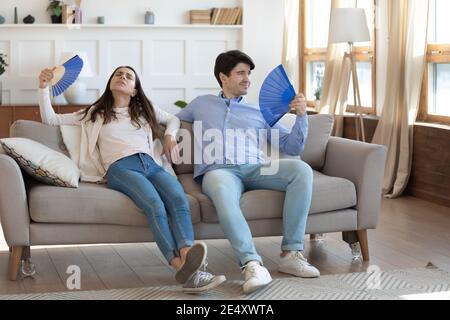 Homme et femme épuisés agitant des ventilateurs de papier, reposant sur un canapé Banque D'Images