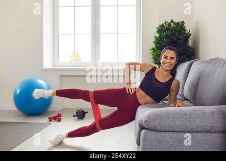 La femme sportive pratique des exercices de résistance à la maison avec des bandes de caoutchouc s'appuyant sur le canapé. Banque D'Images