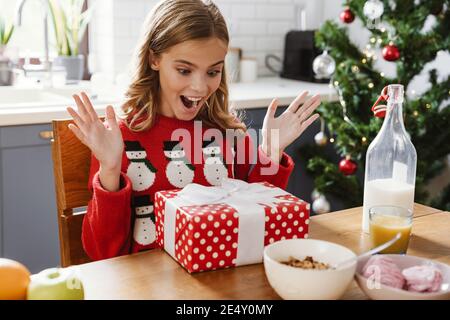 Excitée caucasienne fille ouvrant cadeau de Noël tout en prenant le petit déjeuner à cuisine maison Banque D'Images