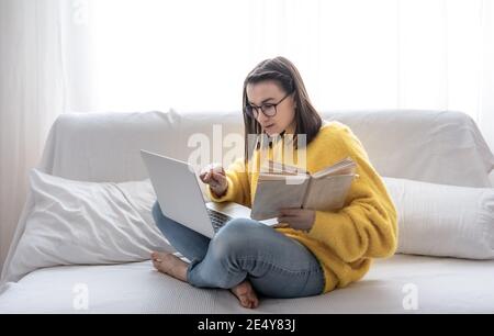 Une fille brune élégante dans un chandail jaune est assise à la maison sur le canapé avec un livre et un ordinateur portable. Le concept de la vie étudiante et de l'auto-développement. Banque D'Images