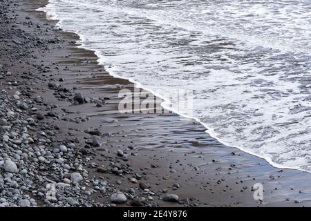 Gros plan sur les vagues de surf arrivant en bord de mer sur le noir vulcanique plage de sable et de rochers Banque D'Images