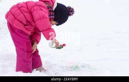 Une jolie fille avec une bouteille de peinture dessine sur la neige. Le concept de la créativité des enfants et du plaisir d'hiver. Banque D'Images