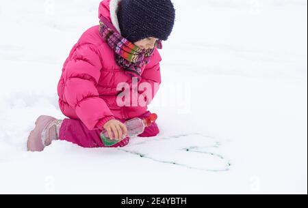 Une jolie fille avec une bouteille de peinture dessine sur la neige. Le concept de la créativité des enfants et du plaisir d'hiver. Banque D'Images