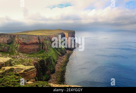 Vue sur la côte d'Orkney avec de hautes falaises rouges et l'océan Atlantique eau sous et nuages blancs sur bleu sk Banque D'Images