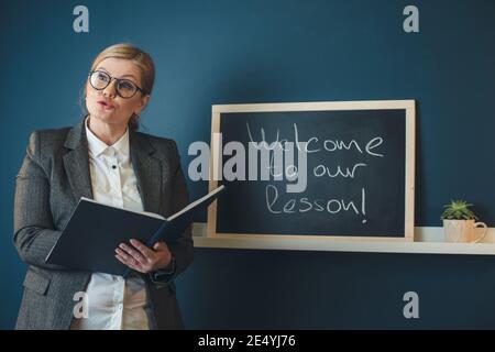 L'enseignant blond senior enseigne devant la classe près du tableau noir tenant un livre Banque D'Images