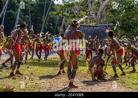 Un passager du navire d'expédition True North se tient dos à la caméra à côté du gardien de wicket accroupi lors d'une partie de cricket de style des îles Trobriand à Kwebwaga, îles Trobriand, Papouasie-Nouvelle-Guinée, avec des danseurs en costume se déplaçant autour du wicket en arrière-plan. Banque D'Images
