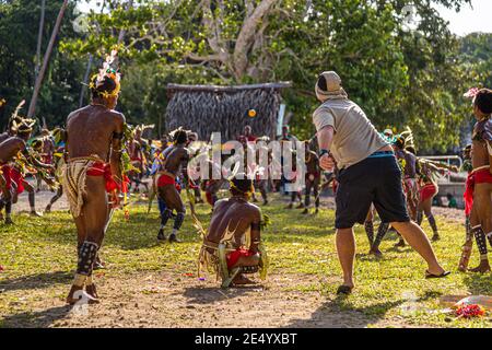 Un passager du navire d'expédition True North lance la balle orange sur le gardien de wicket accroupi lors d'une partie de cricket de style des îles Trobriand à Kwebwaga, îles Trobriand, Papouasie-Nouvelle-Guinée, un danseur costumé se tenant à ses côtés, d'autres danseurs à l'arrière-plan devant une hutte en chaume de palmiers. Banque D'Images