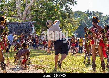 Un passager du navire d'expédition True North lance la balle orange lors d'une partie de cricket style îles Trobriand à Kwebwaga, îles Trobriand, Papouasie-Nouvelle-Guinée, vêtu d'une tenue décontractée et d'un couvre-chef, un danseur costumé accroupi devant le guichet, des hommes costumés debout autour. Banque D'Images