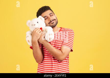 Un homme heureux satisfait avec une barbe en t-shirt rayé qui embrasse un adorable ours blanc en peluche, qui ferme les yeux et rêve, en appréciant un cadeau romantique. Prise de vue en studio I Banque D'Images