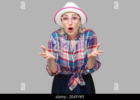 Portrait de femme mature élégant moderne surpris dans un style décontracté avec chapeau et lunettes debout, bras levés, looking at camera avec choqué face. Banque D'Images