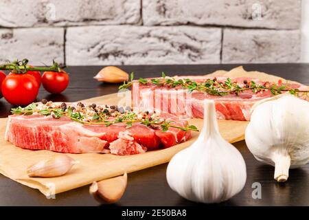 steaks de porc crus fraîchement hachés avec épices et thym sur une feuille de parchemin, à côté de tomates rouges mûres et d'ail Banque D'Images