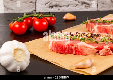 steaks de porc crus fraîchement hachés avec épices et thym sur une feuille de parchemin, à côté de tomates rouges mûres et d'ail Banque D'Images