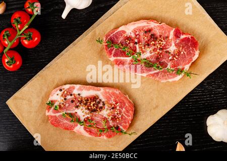 steaks de porc crus fraîchement hachés avec épices et thym sur une feuille de parchemin, à côté de tomates rouges mûres et d'ail Banque D'Images