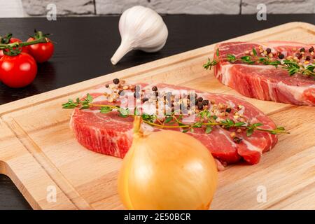 steaks de porc frais hachés avec épices et thym sur une planche à découper, à côté de tomates rouges mûres et d'ail Banque D'Images