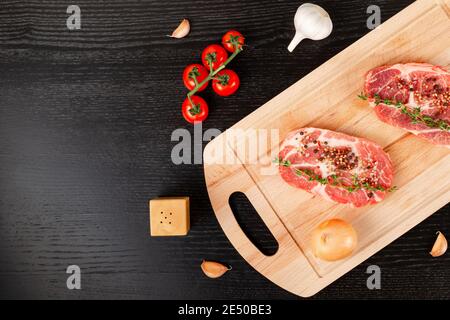 steaks de porc frais hachés avec épices et thym sur une planche à découper, à côté de tomates rouges mûres et d'ail Banque D'Images