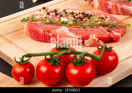 steaks de porc frais hachés avec assaisonnement, tomates mûres et thym sur une planche à découper sur une table en bois noir Banque D'Images