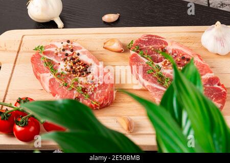 steaks de porc frais hachés avec épices et thym sur une planche à découper, à côté de tomates rouges mûres et d'ail Banque D'Images